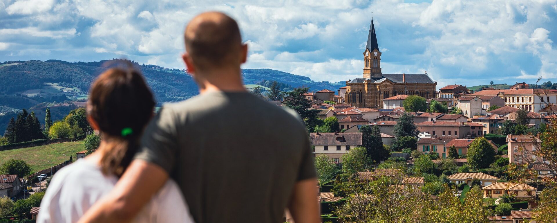Un couple de dos qui regarde le village de Bessenay. Vue sur son église et les Monts du Lyonnais.