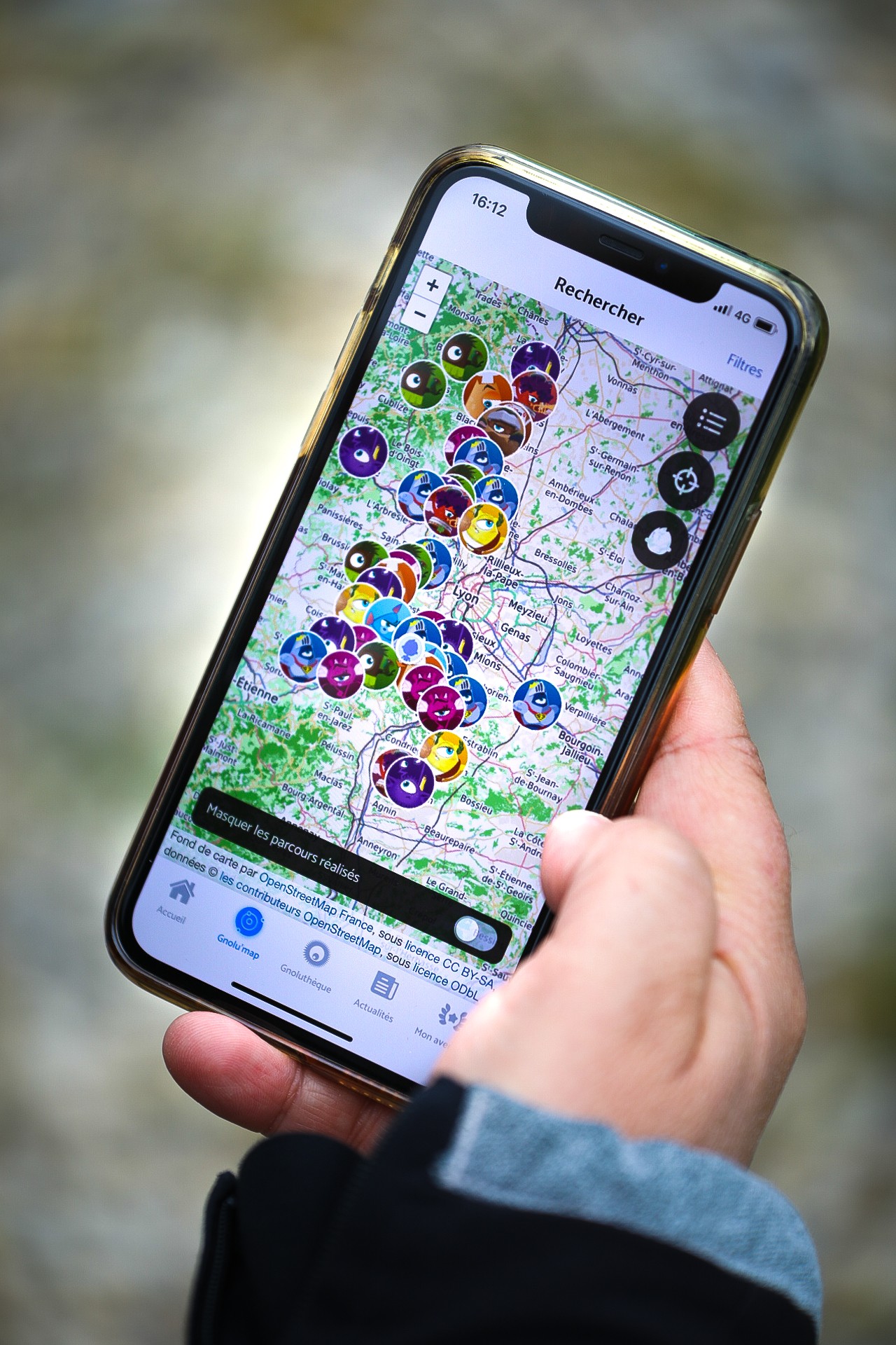 Zoom sur une main tenant un téléphone avec l'application Les Gnolus ouverte. Cette application permet de découvrir le département du Rhône de façon ludique. Présentation des lieux de caches sur l'application.
