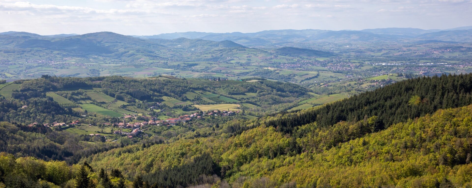 Vue sur les Monts du Lyonnais avec au loin, le village de Pomeys. Ce petit village est entouré de forêts et champs dans un paysage vallonné.