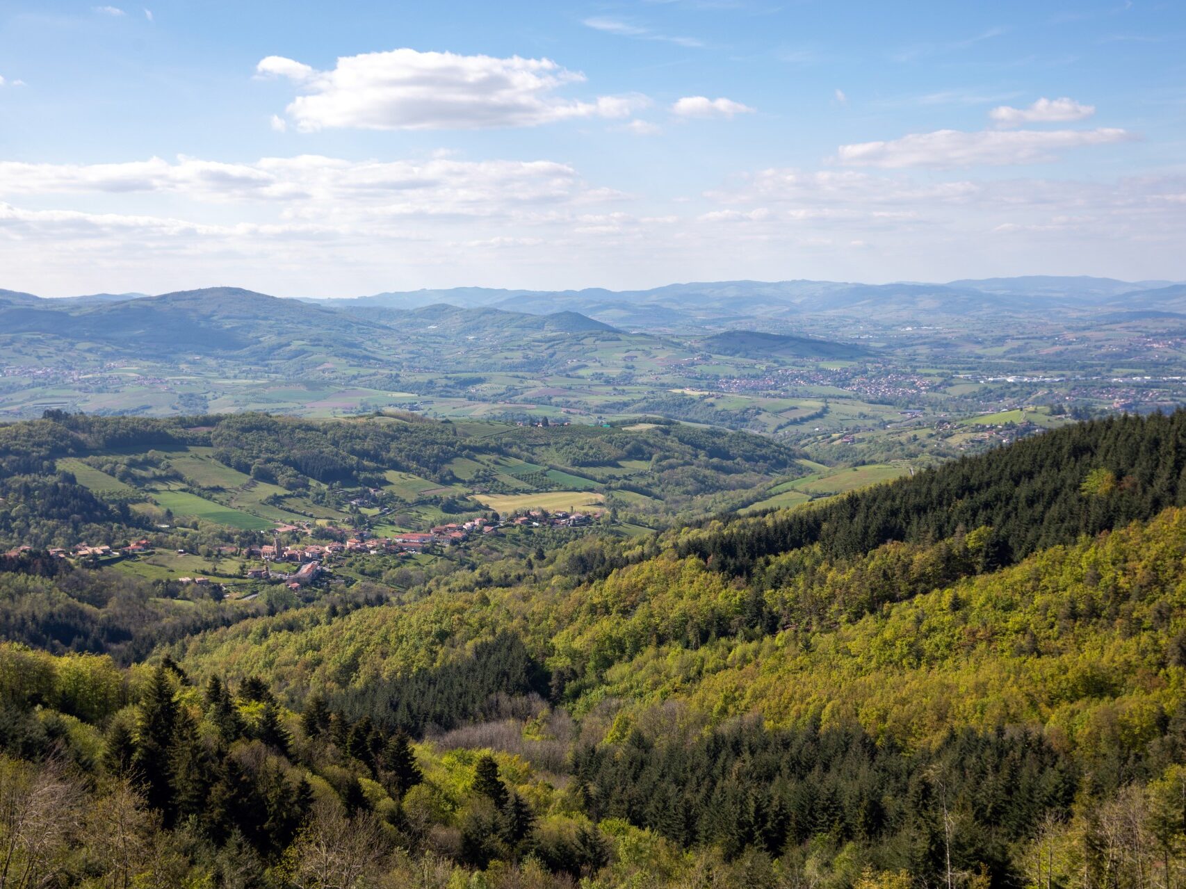 Vue sur les Monts du Lyonnais avec au loin, le village de Pomeys. Ce petit village est entouré de forêts et champs dans un paysage vallonné.