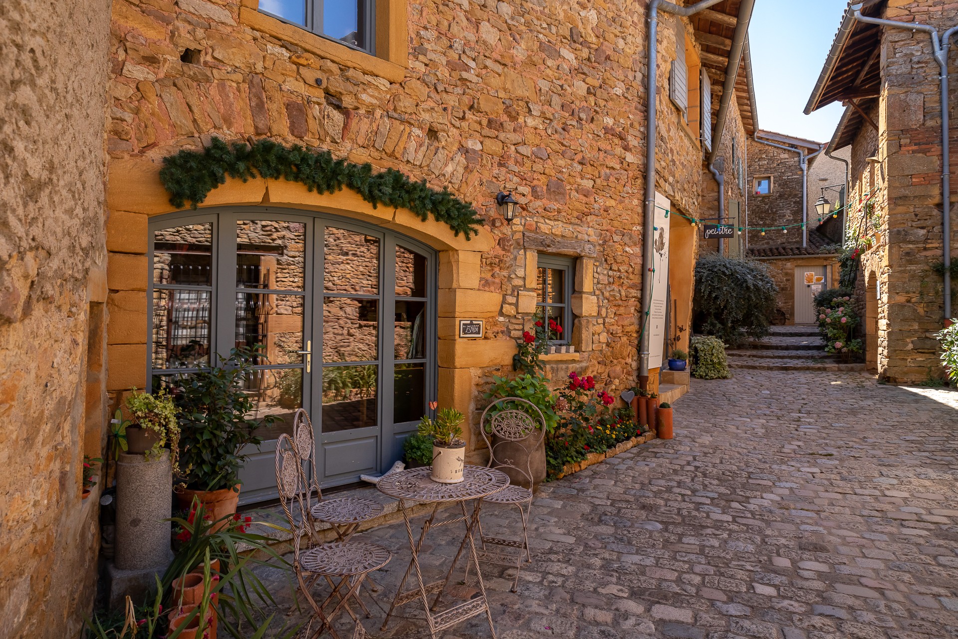 Village d'Oingt, ruelle pavée entourées de maisons en pierres dorées en été. Petit salon de jardin en fer forgé au premier plan