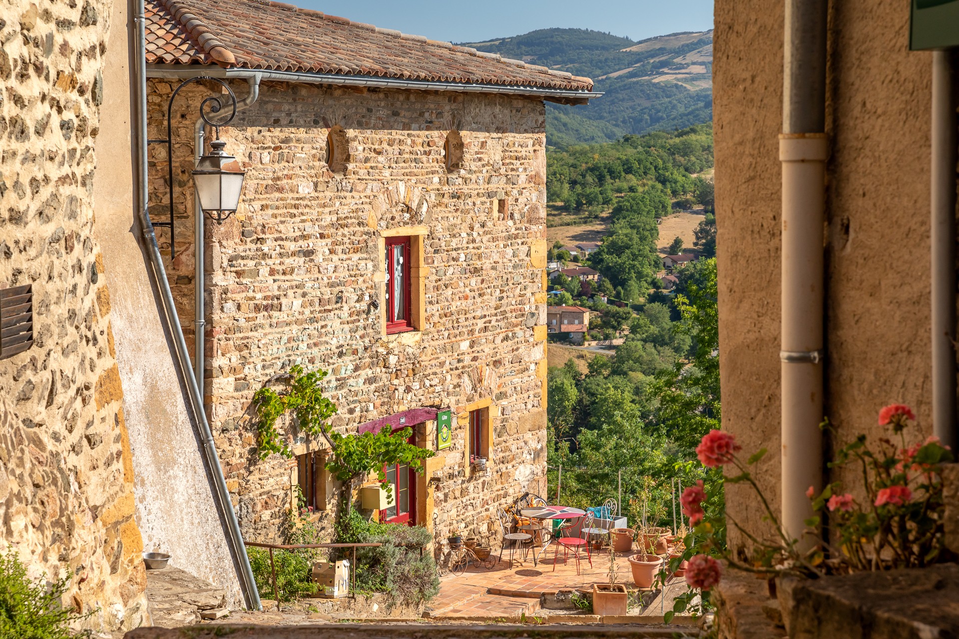 Vue plongeante sur une rue pavée du vieux bourg médiéval de Ternand. En contre-bas de la rue, un salon de jardin en fer forgé est installé au pied d'une maison en pierres apparentes. Colline verdoyante en en arrière plan. Beaujolais en été