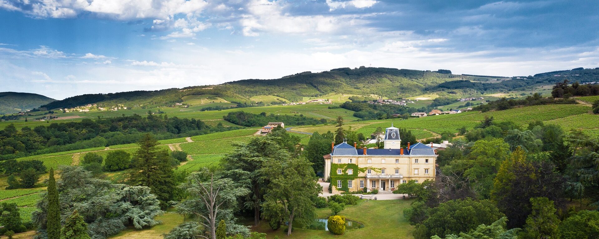 Vue drone sur le Château de Bellevue, son parc arboré et verdoyant. Vignoble et collines boisées du Haut-Beaujolais du Beaujolais en arrière-plan. Ciel nuageux et bleu.