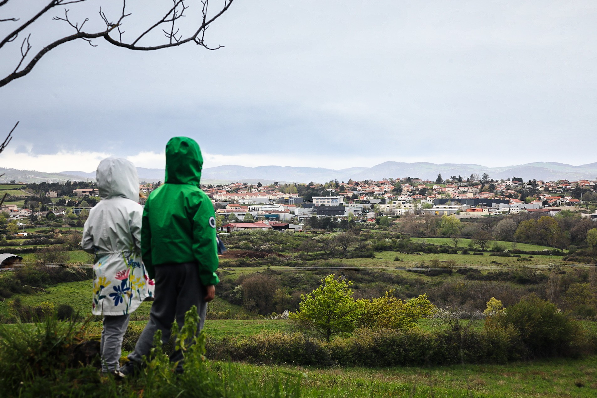 2 enfants de dos avec un imperméable et leur capuche regardent les Monts du Lyonnais et un village.