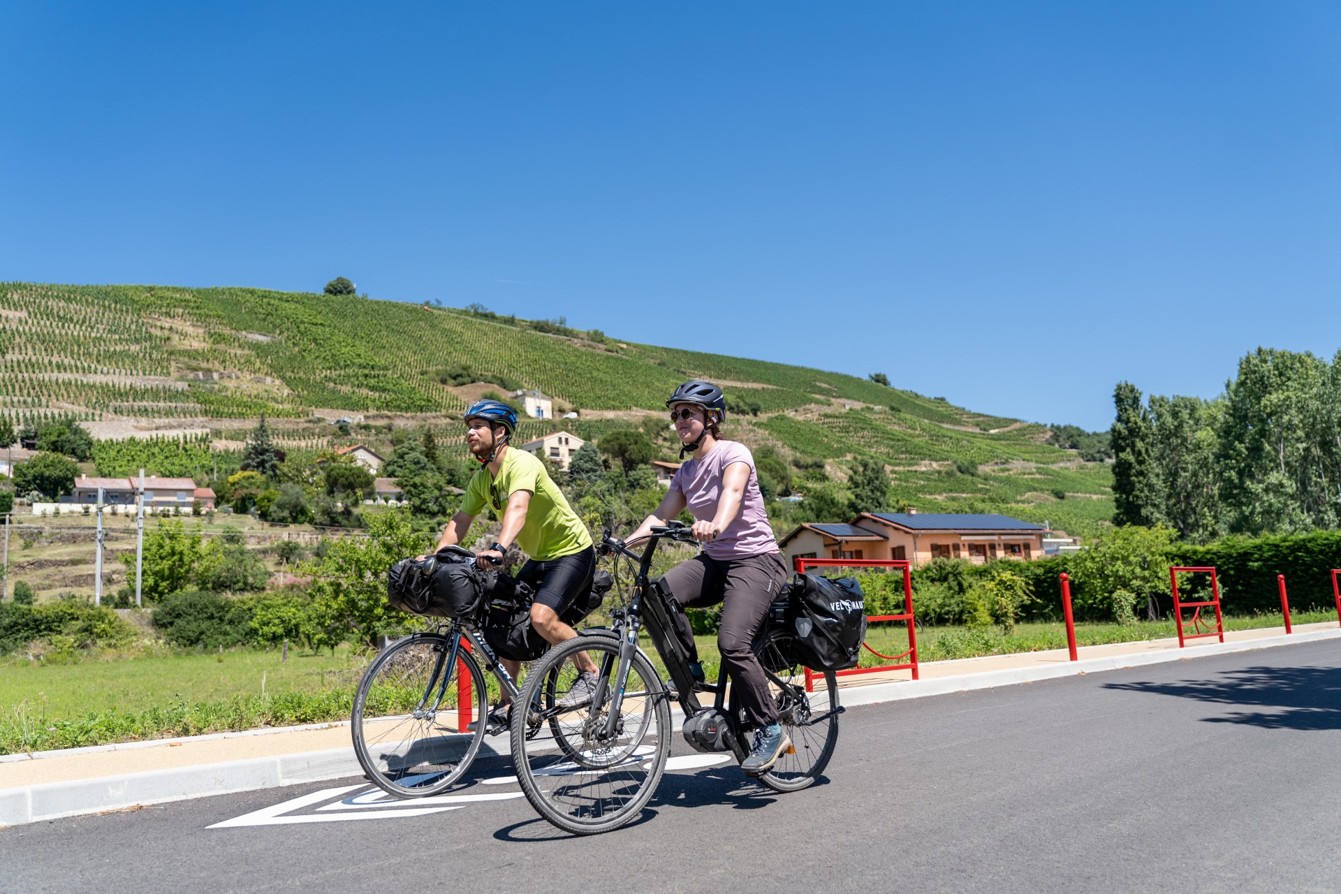 Un couple d'itinérants roulant côte à côte à vélo avec leur bagages sur la ViaRhôna à Ampuis avec une vue sur les côteaux de la Cote-Rôtie tous proches.