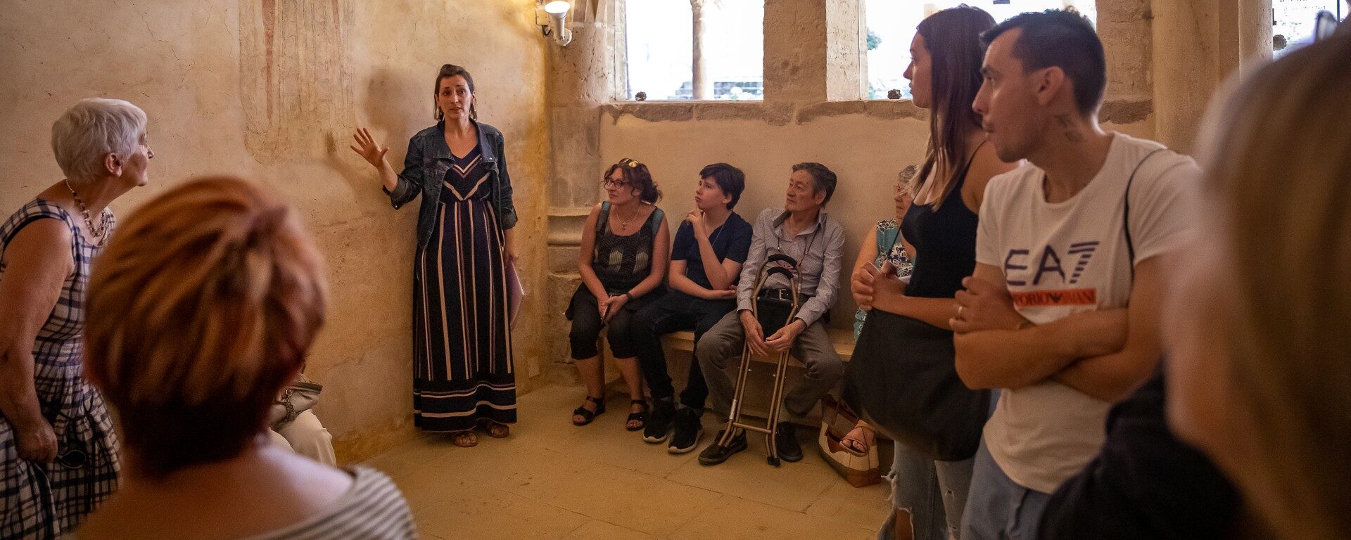 Groupe en visite guidée dans le cloître du prieuré roman de Salles dans le Beaujolais. La guide présente la fresque murale représentant Saint-Odilon dans la salle capitulaire