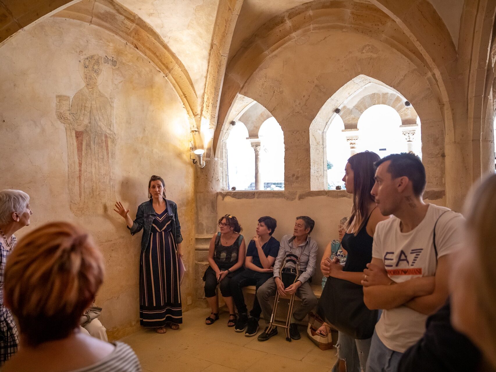Groupe en visite guidée dans le cloître du prieuré roman de Salles dans le Beaujolais. La guide présente la fresque murale représentant Saint-Odilon dans la salle capitulaire
