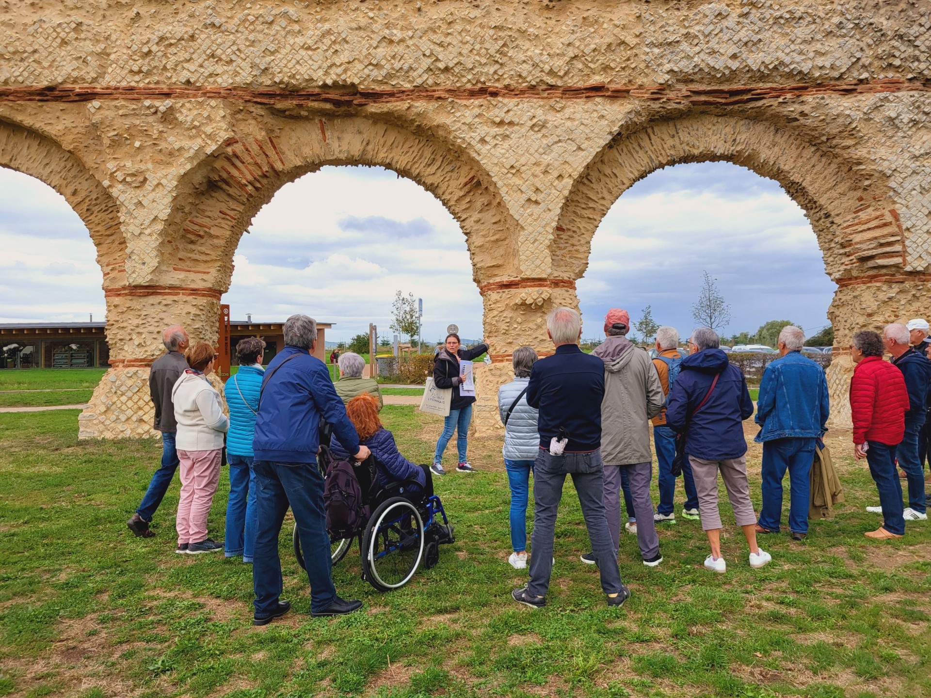 Un groupe de personnes participe à une visite guidée en plein air devant un ancien aqueduc en pierre. La guide explique des détails historiques pendant que les visiteurs l'écoutent attentivement. Une personne en fauteuil roulant est présente, mettant en avant l'inclusivité du groupe.