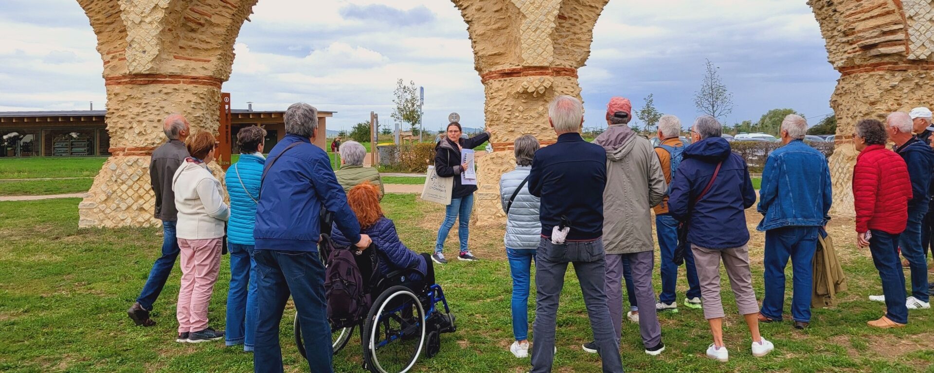 Un groupe de personnes participe à une visite guidée en plein air devant un ancien aqueduc en pierre. La guide explique des détails historiques pendant que les visiteurs l'écoutent attentivement. Une personne en fauteuil roulant est présente, mettant en avant l'inclusivité du groupe.
